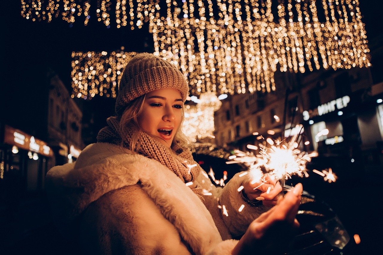 O femeie tânără bucurându-se de sărbătorile de iarnă pe o stradă ornamental iluminată, ținând în mână o bengală scânteietoare, cu zâmbetul strălucitor de bucurie și uimire înconjurate de lumini festive.
