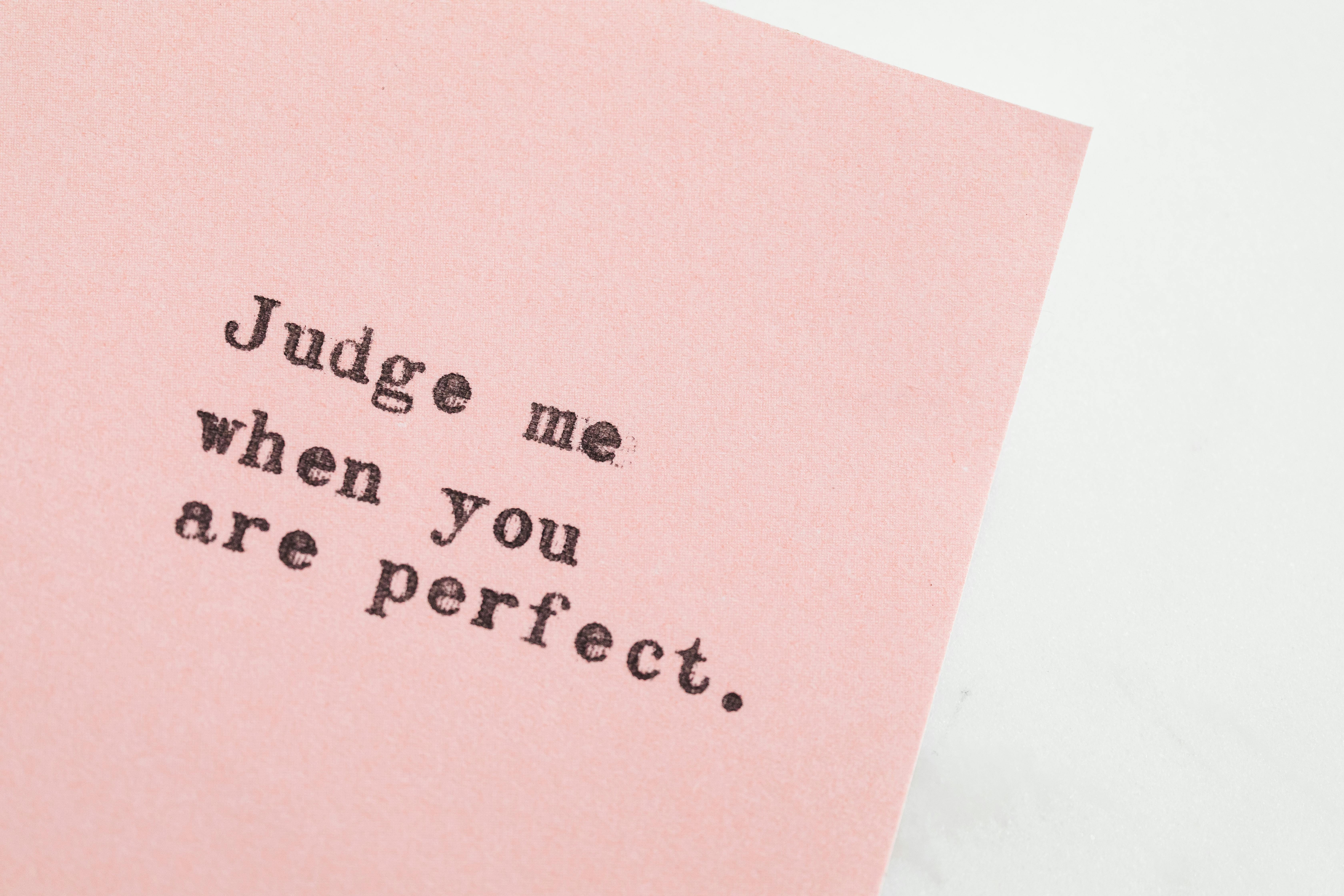 Imagine cu mesaj tipărit pe o foaie roz deschis: 'Judge me when you are perfect'. Textul subliniază tema perfecționismului și a presiunii de a fi perfect.